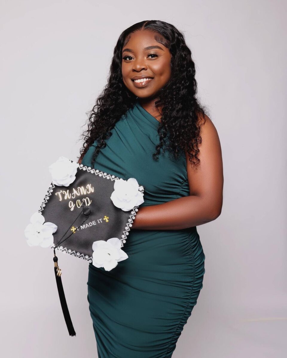 A Woman In A Green Dress Holds A Decorated Graduation Cap With The Words &Quot;Thank God I Made It&Quot; And White Flower Embellishments, Smiling Proudly Against A Plain Background—A Moment Celebrated By Red October Firm.