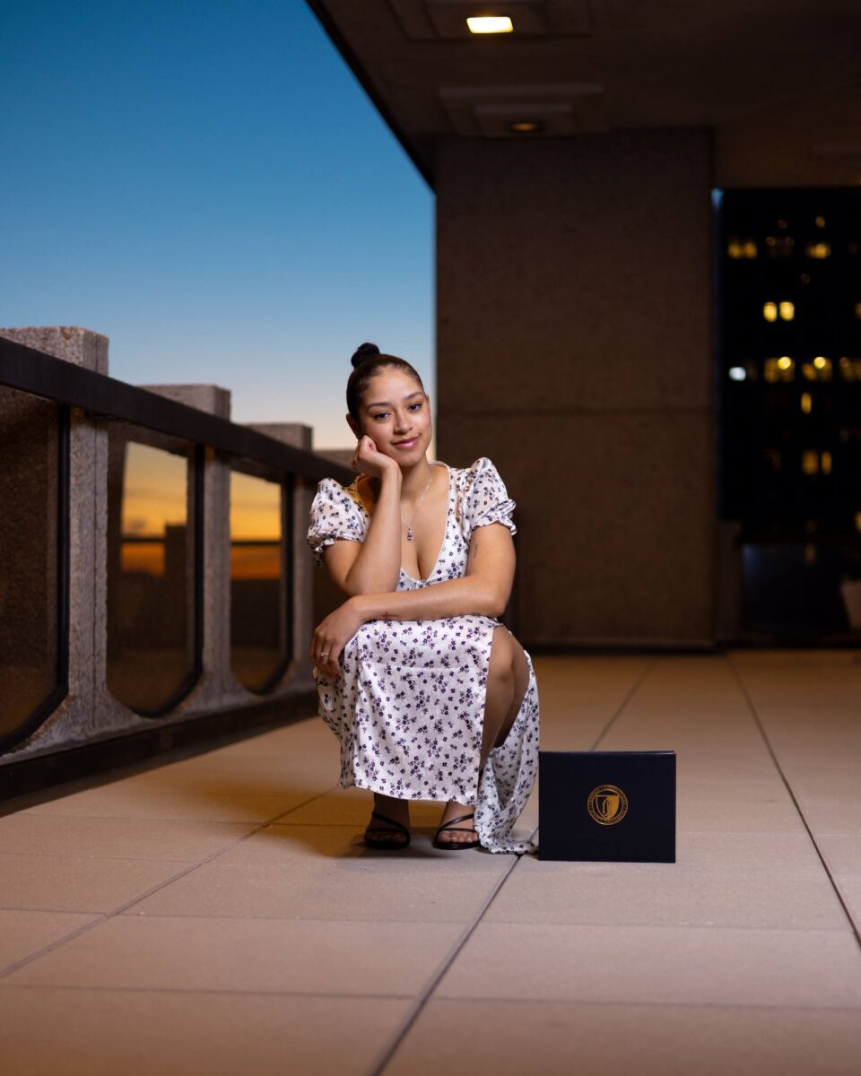 A Woman In A White Floral Dress Squats On A Balcony At Sunset, Resting Her Chin On Her Hand Beside A Black Folder With The Red October Firm Gold Emblem.
