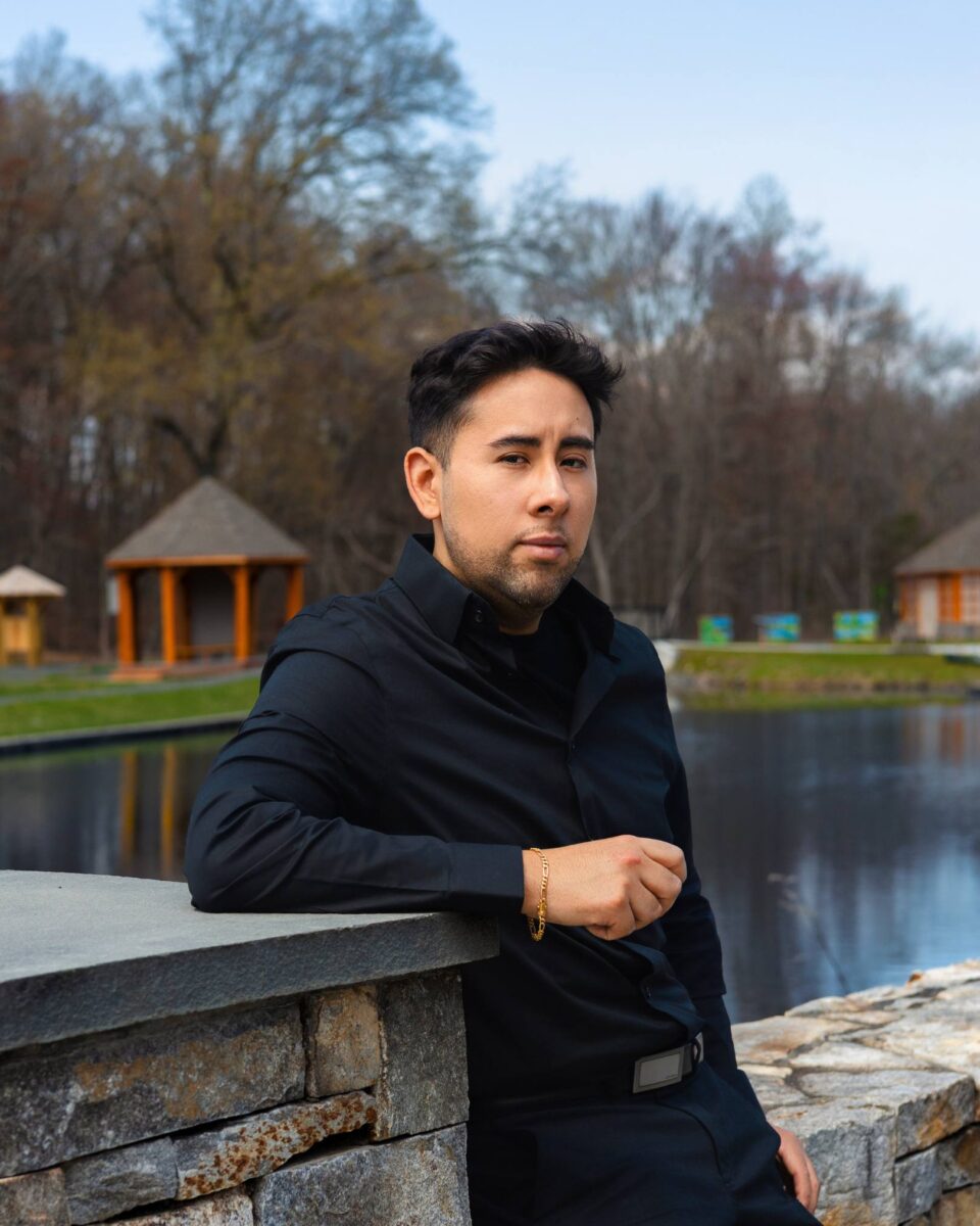 A Man In A Black Shirt Leans Against A Stone Wall Near A Pond With Wooden Gazebos And Trees In The Background, Embodying The Calm Confidence Of Red October Firm.