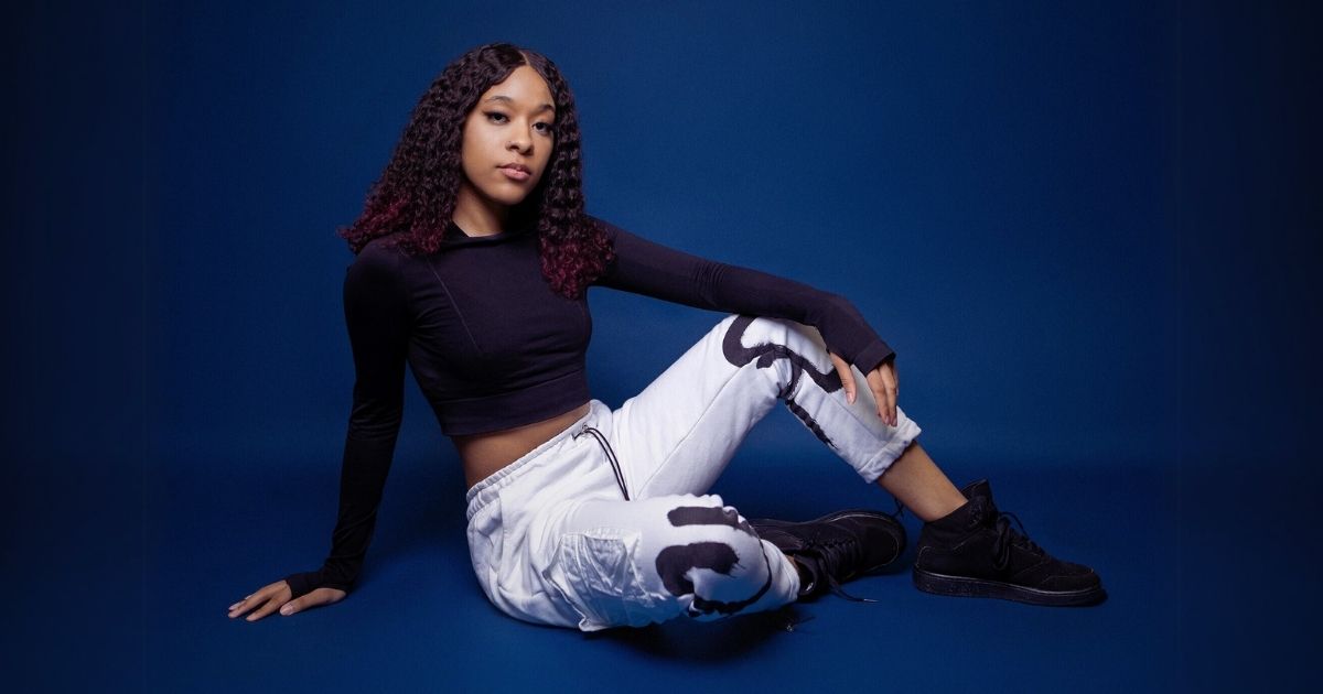 A Woman Posing In A Black Top And White Pants On A Blue Background.