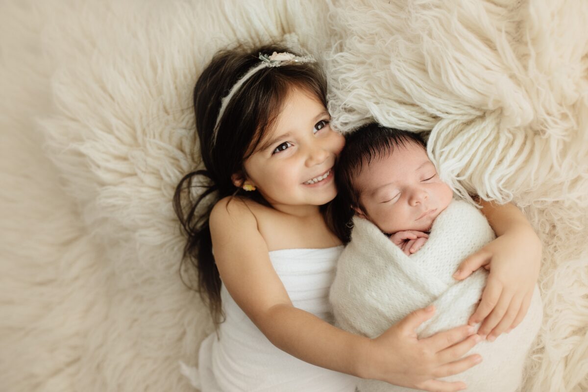 A Little Girl Poses With Her Newborn, Wrapped Up In A Blanket, During A Heartwarming Photo Session.