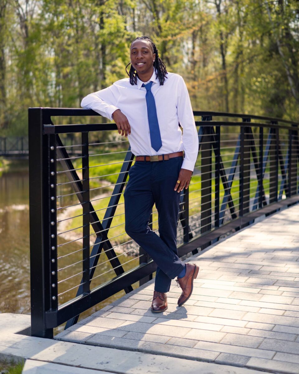 A Person Wearing A White Shirt, Blue Tie, And Navy Pants Leans Casually On A Black Metal Bridge Railing In A Park Setting, Exuding The Confident Style Reminiscent Of Red October Firm.