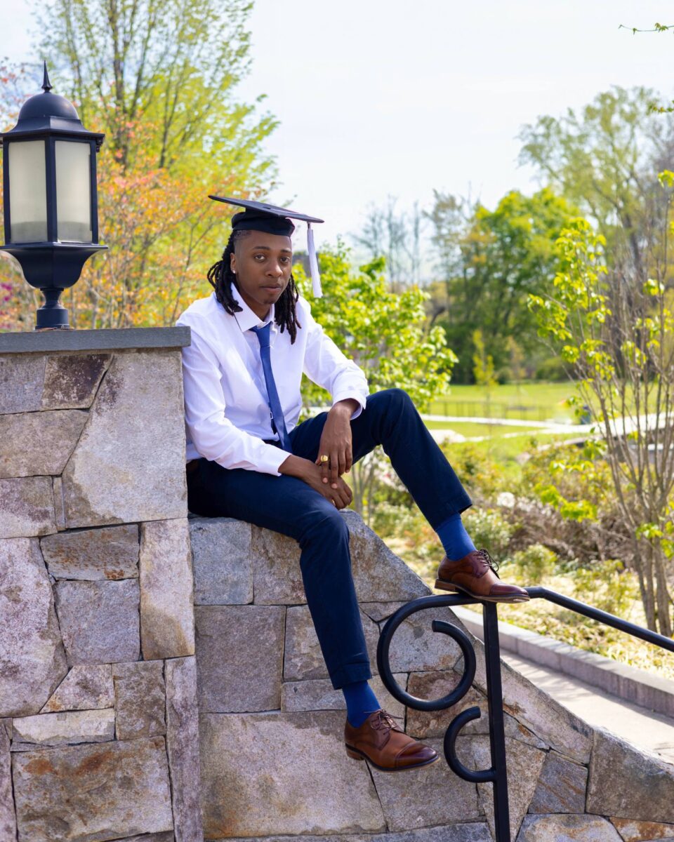 A Young Man Wearing A Graduation Cap, White Shirt, And Tie Sits On A Stone Wall Outdoors—An Inspiring Moment Captured By Red October Firm Amid Greenery And A Lamp Post In The Background.