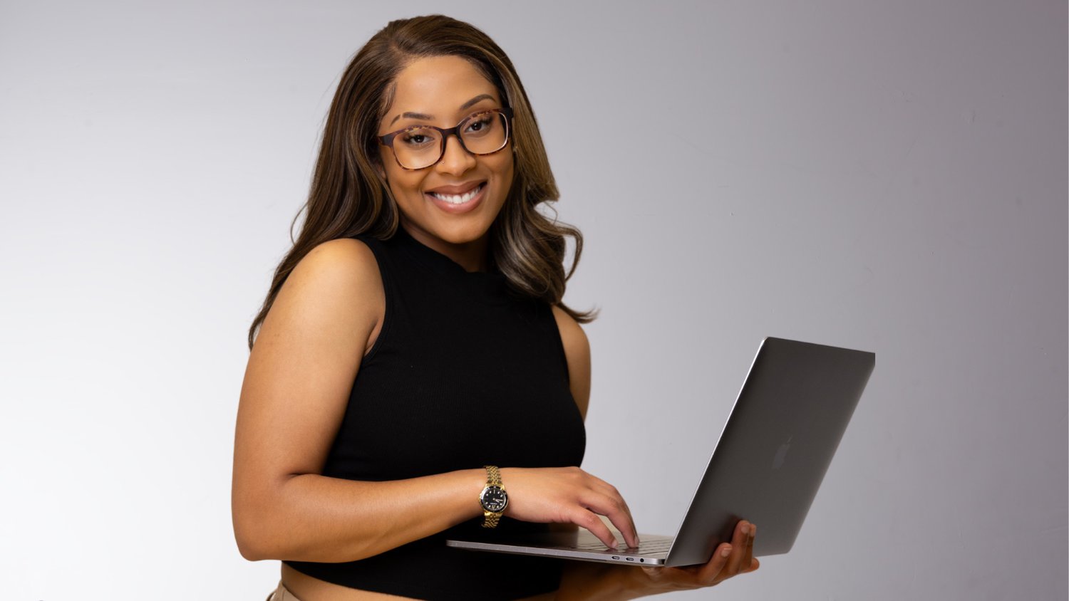 A Woman With Glasses, Wearing A Black Sleeveless Top, Smiles At The Camera While Holding A Laptop, Ready To Offer Her Services.