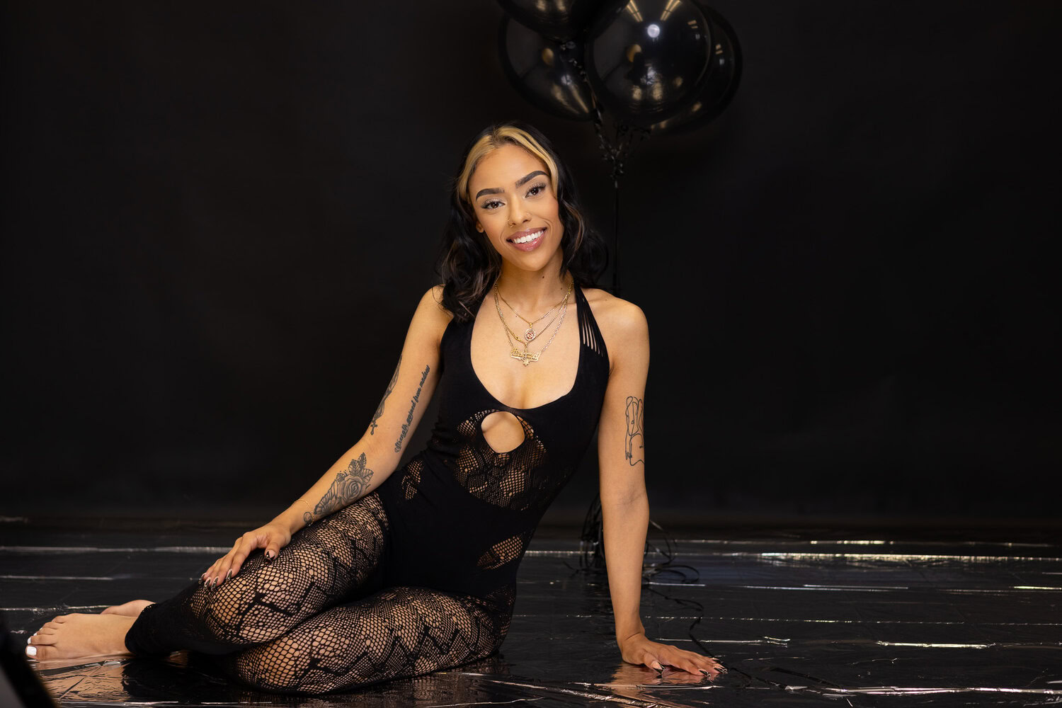 A Woman With Tattoos, Wearing A Black Patterned Outfit, Sits On The Floor Against A Dark Backdrop With Black Balloons In The Background, Offering Her Unique Services.