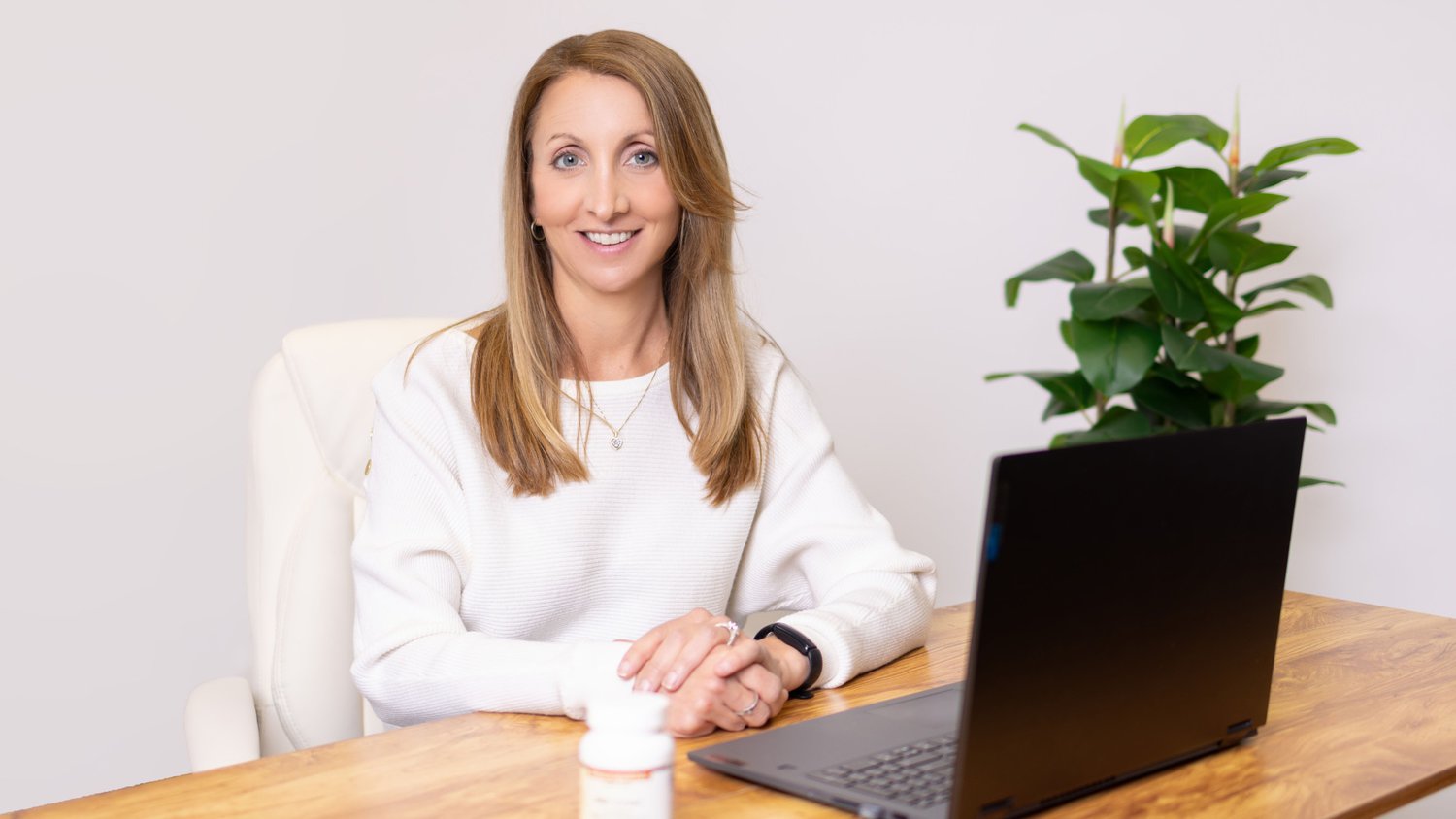 A Woman In A White Sweater Sits At A Desk With A Laptop And A Medication Bottle, Possibly Reviewing Healthcare Services. A Green Plant Is In The Background.