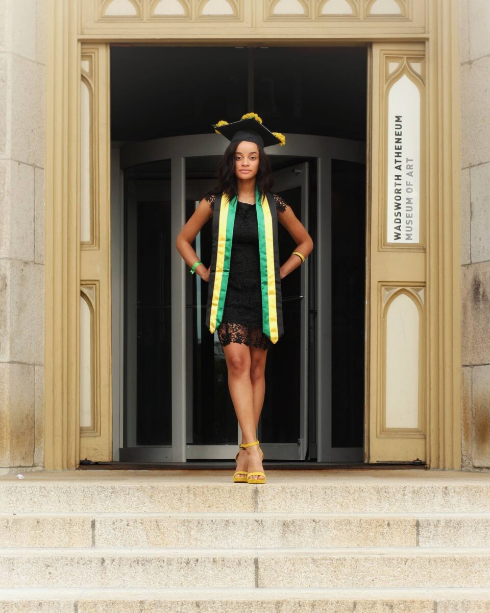 A Woman In A Graduation Cap And Gown Stands On Steps In Front Of A Building Labeled &Quot;Wadsworth Atheneum Museum Of Art,&Quot; Her Achievement Celebrated By Red October Firm.